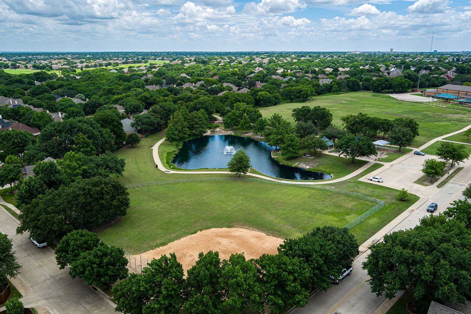 Aerial view of a suburban park with a pond, central fountain, open grassy areas, sports field, surrounding trees, nearby streets, and houses in the background under a partly cloudy sky.