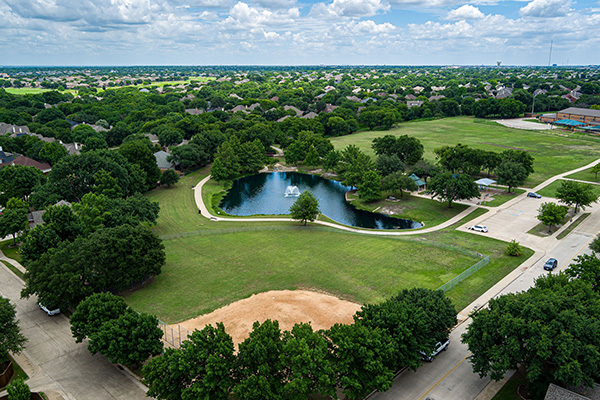 Aerial view of a suburban park with open grassy areas, a pond with a fountain, surrounding trees, and houses in the background under a partly cloudy sky.