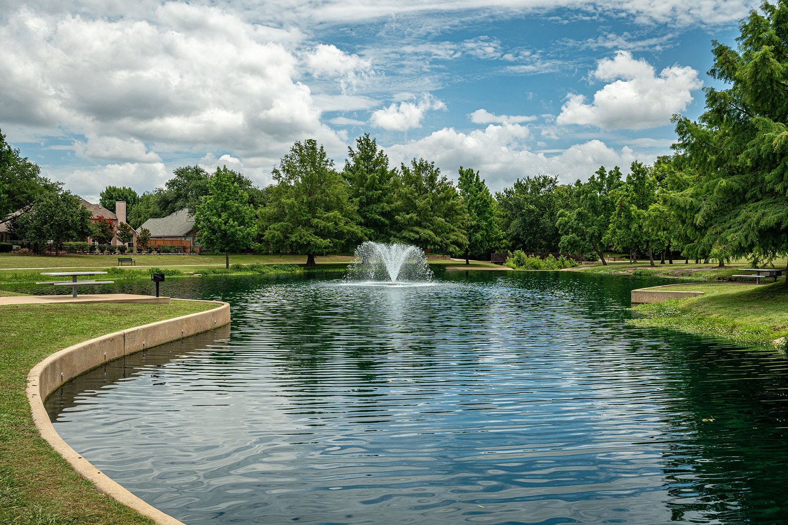 A small park pond with a central water fountain, surrounded by green grass and trees, under a partly cloudy sky. Benches and a house are visible in the background.