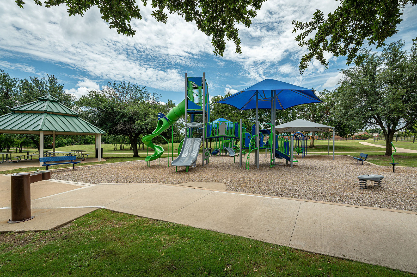 A playground with slides and climbing structures sits on mulch, surrounded by trees, benches, and covered picnic areas under a partly cloudy sky.