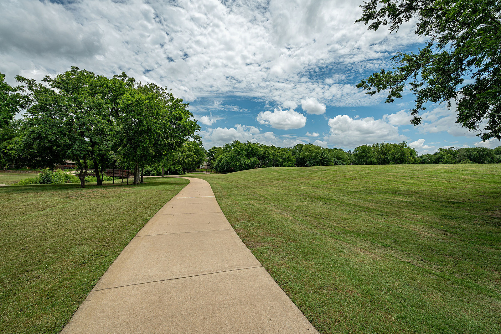 A concrete path curves through a grassy park with scattered trees under a partly cloudy sky.