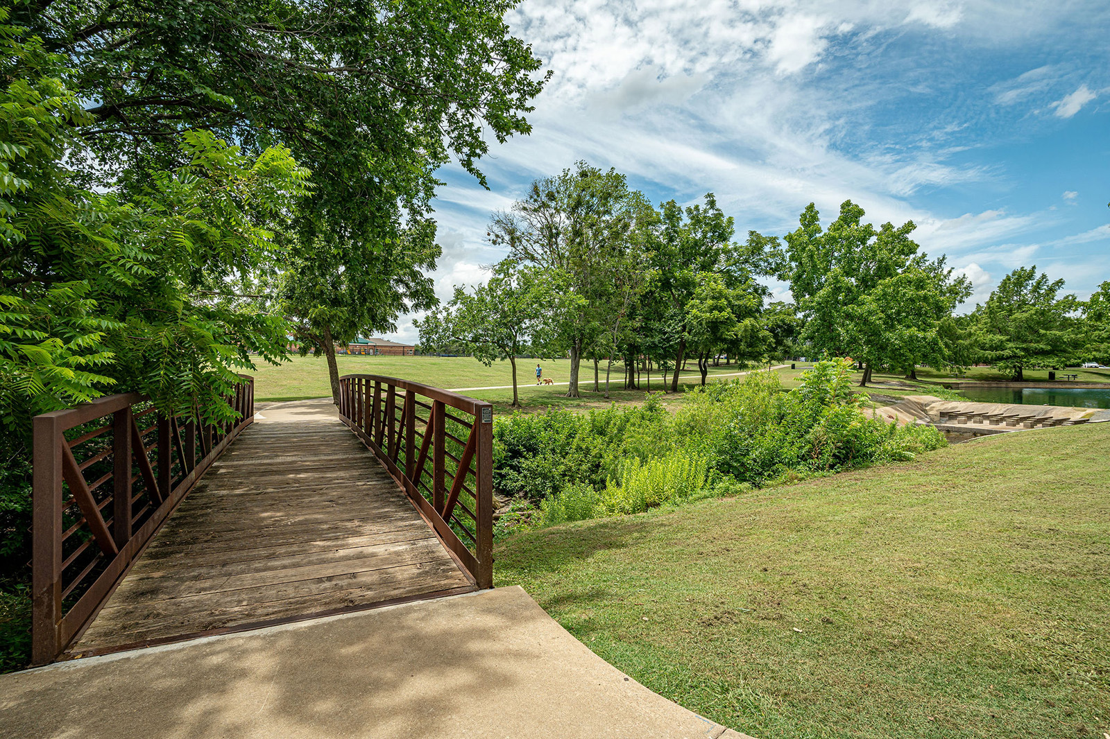 A wooden footbridge crosses a grassy area in a park with green trees, a clear sky, and a person visible in the distance.