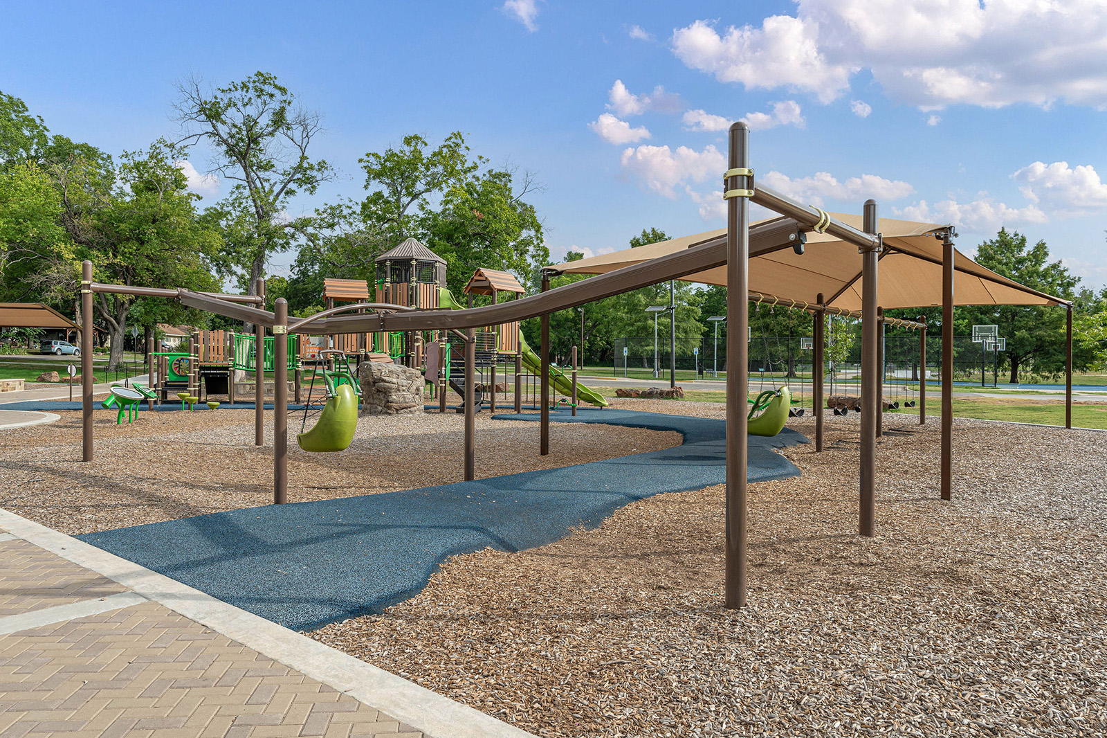 Modern playground with climbing structures, slides, shaded areas, and wood chip ground cover, surrounded by trees and open sky.