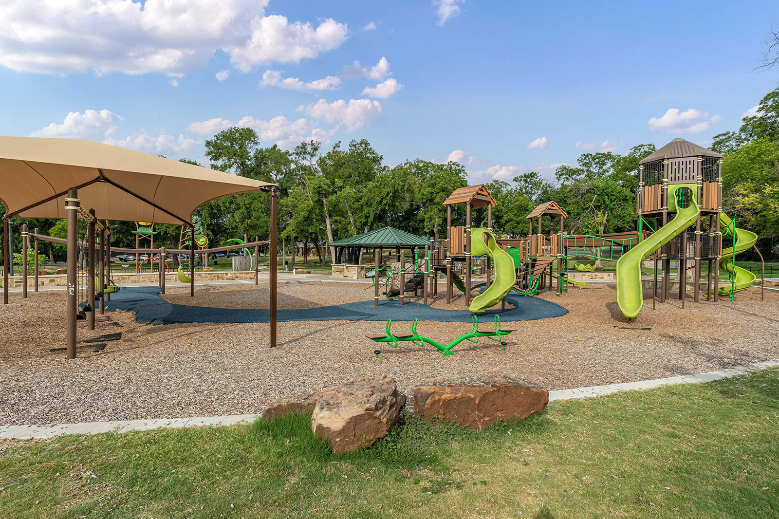 Outdoor playground with green slides, climbing structures, and swings on a gravel surface. Shade canopy and trees are in the background under a blue sky with scattered clouds.