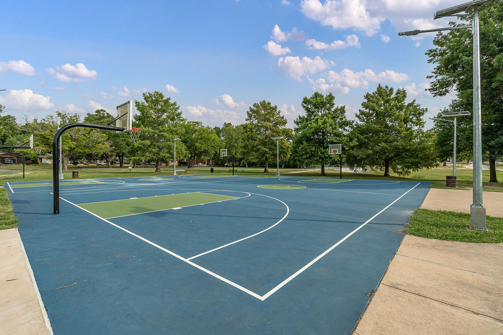Outdoor basketball court with blue and green surface, surrounded by trees and light poles, under a partly cloudy sky.