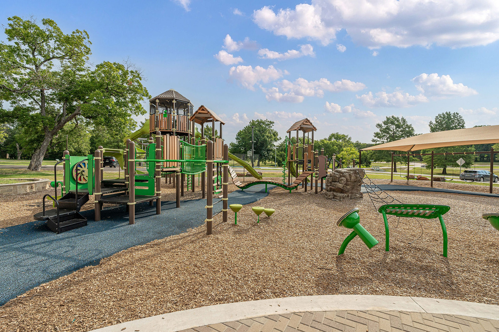 Playground with green and brown climbing structures, slides, and swings on mulch and rubber surfacing; trees and covered picnic area in the background under a partly cloudy sky.