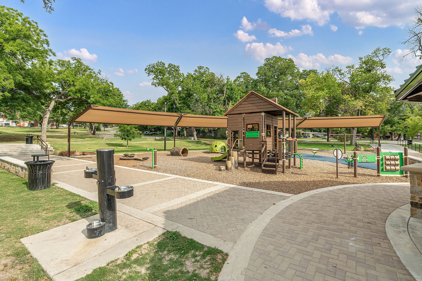 A shaded playground with a wooden play structure, slides, climbing features, and surrounding benches in a park setting with trees and a paved walkway.