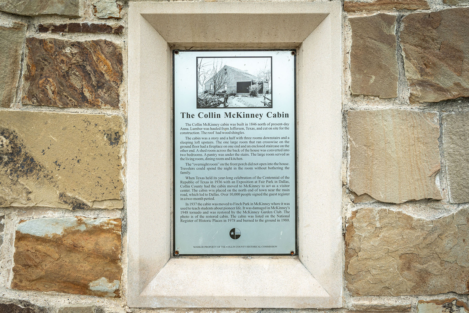 A stone wall features a framed plaque titled "The Collin McKinney Cabin," with historical information and a black-and-white cabin photo at the top.