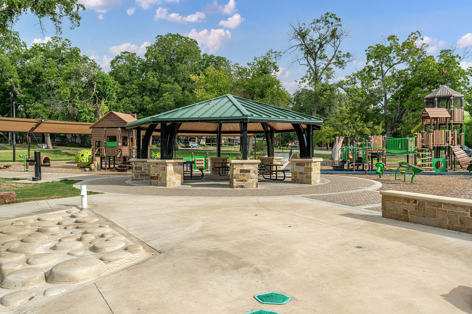 A covered pavilion with picnic tables stands in the center of a park, surrounded by playground equipment, trees, and open space under a partly cloudy sky.