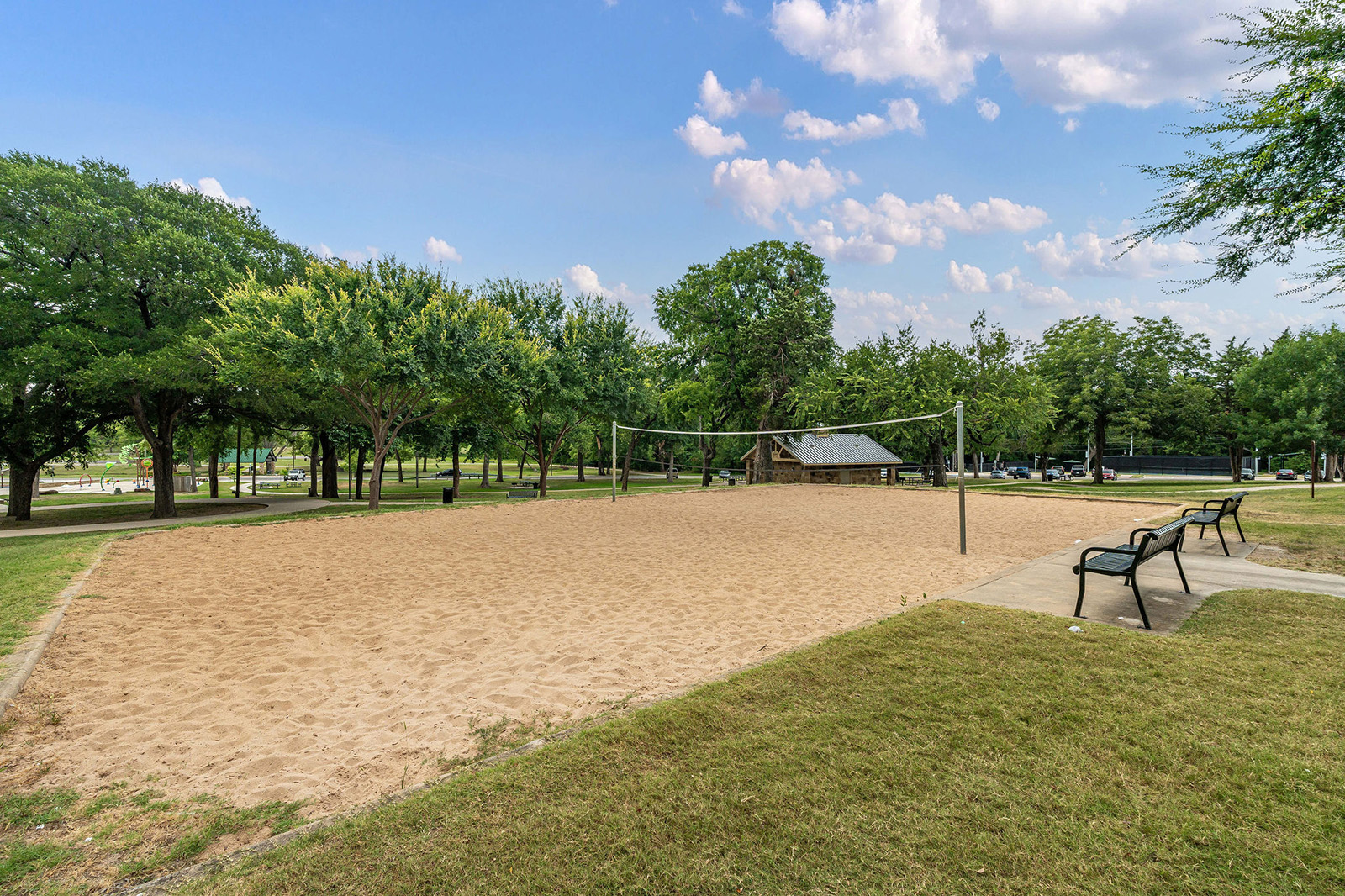 An empty sand volleyball court in a park, surrounded by trees, benches, and a small pavilion under a partly cloudy sky.