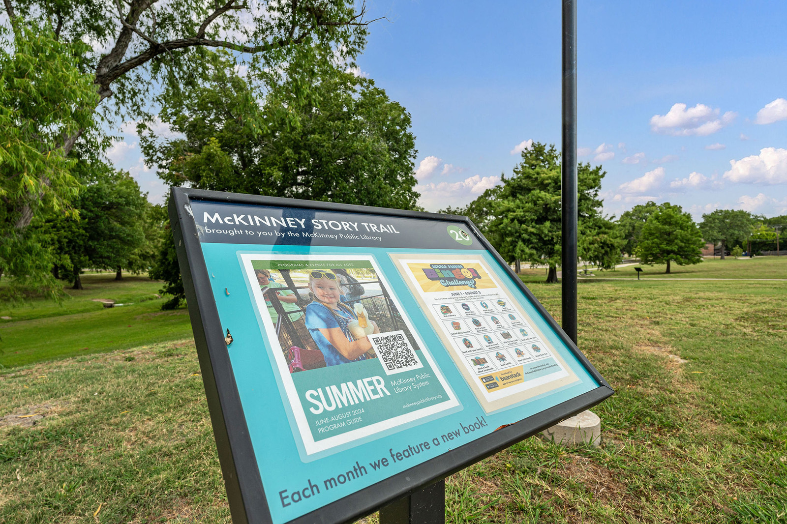 Outdoor sign for the McKinney Story Trail in a grassy park, featuring information about a summer reading program and a monthly featured book.