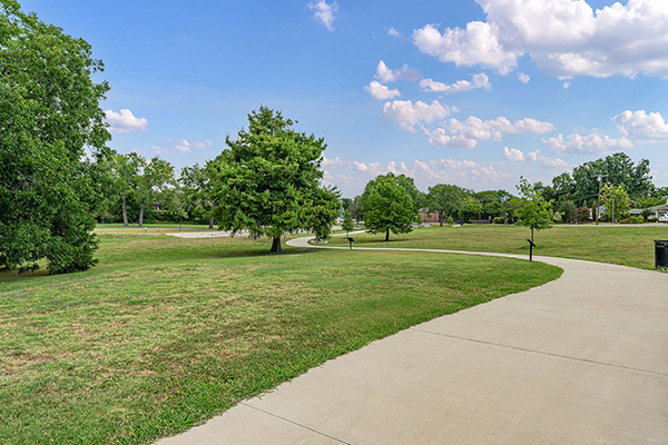 A paved walking path curves through a grassy park with scattered trees under a partly cloudy sky.