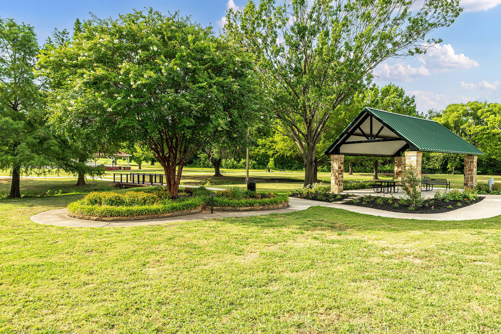 A park with green grass, trees, a picnic shelter with a green roof, picnic tables, and a sunny, partly cloudy sky.