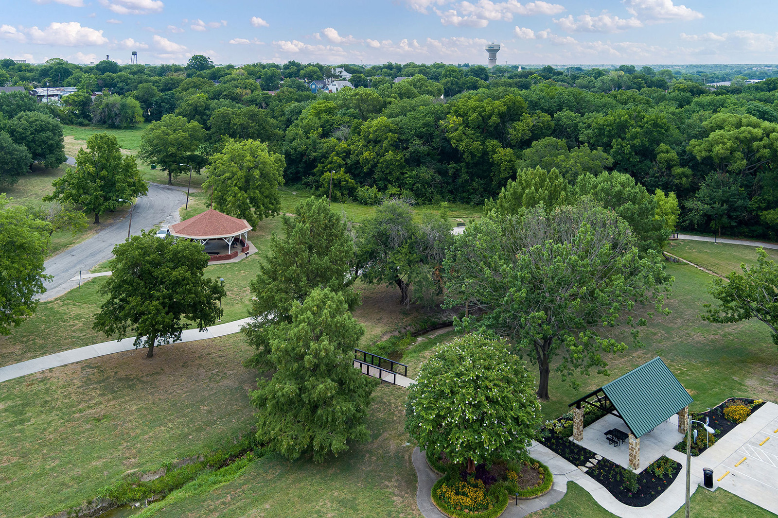 Aerial view of a park with green trees, walking paths, a gazebo, a pavilion, and landscaped flower beds on a partly cloudy day.