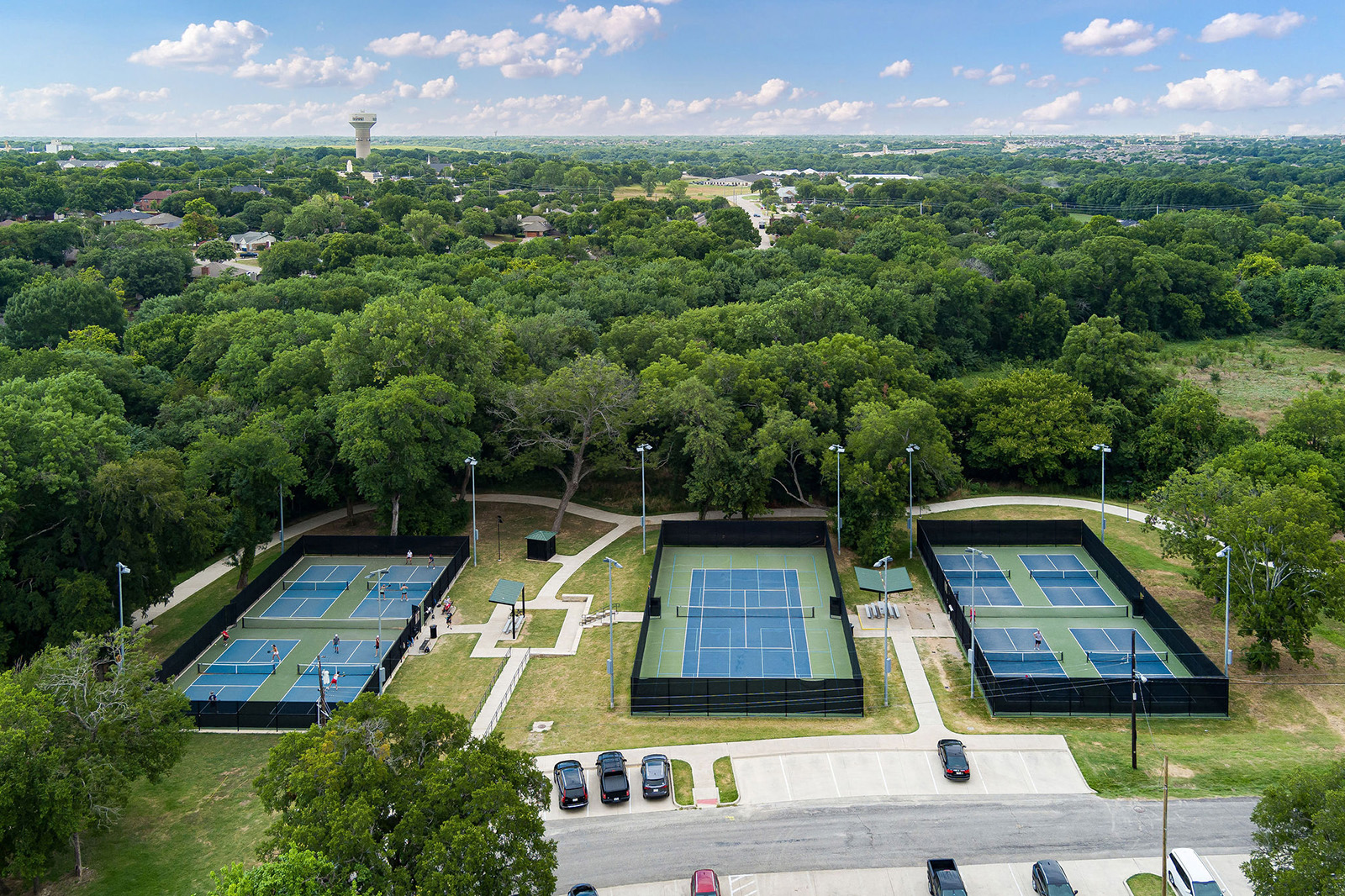 Aerial view of an outdoor sports complex with multiple tennis and pickleball courts, surrounded by trees, parking lot, and a water tower in the distance.