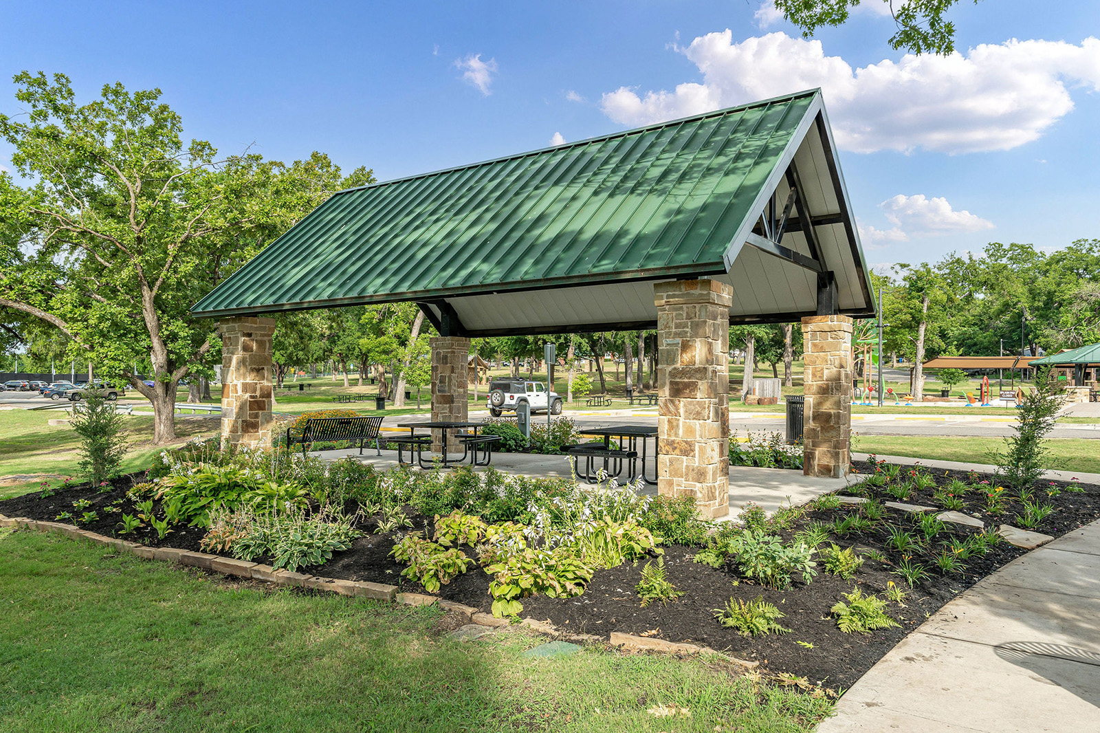 A covered pavilion with a green metal roof and stone pillars is surrounded by landscaped greenery and several picnic tables in a public park.