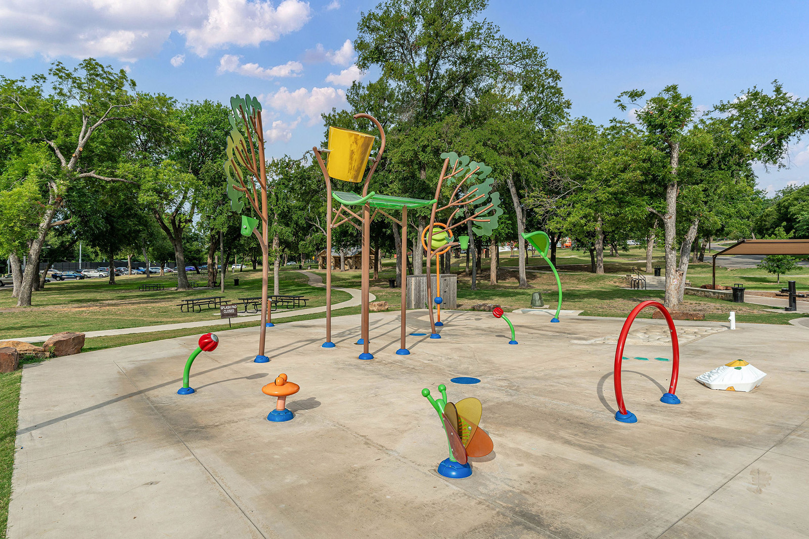 An outdoor splash pad with colorful water features, including a large bucket, flower and leaf designs, set on concrete with trees and picnic tables in the background.