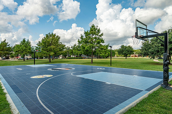 Outdoor basketball court with two hoops, surrounded by grass and trees under a partly cloudy sky.