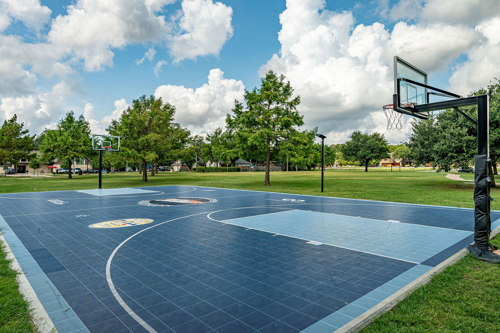 Outdoor basketball court with blue and black tiles, two hoops, and surrounding green grass and trees under a partly cloudy sky.