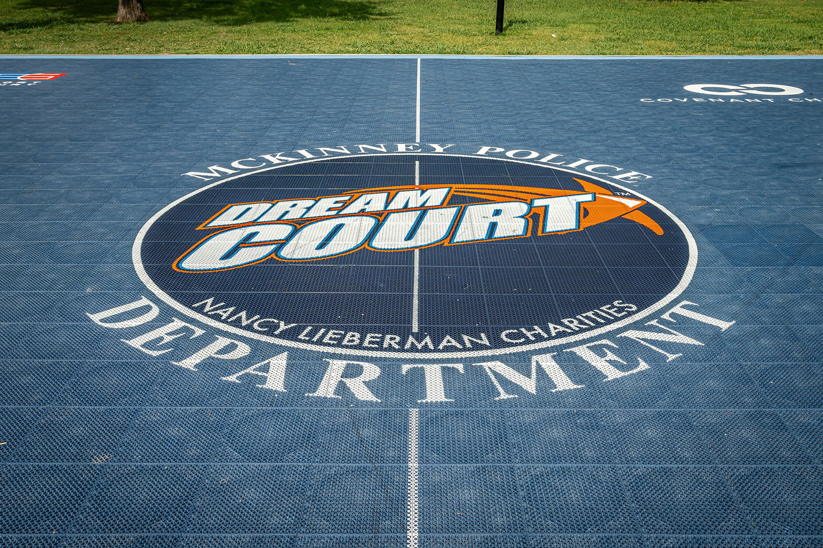 Blue outdoor basketball court labeled "Dream Court," with "McKinney Police Department" and "Nancy Lieberman Charities" printed around a basketball graphic at center court.