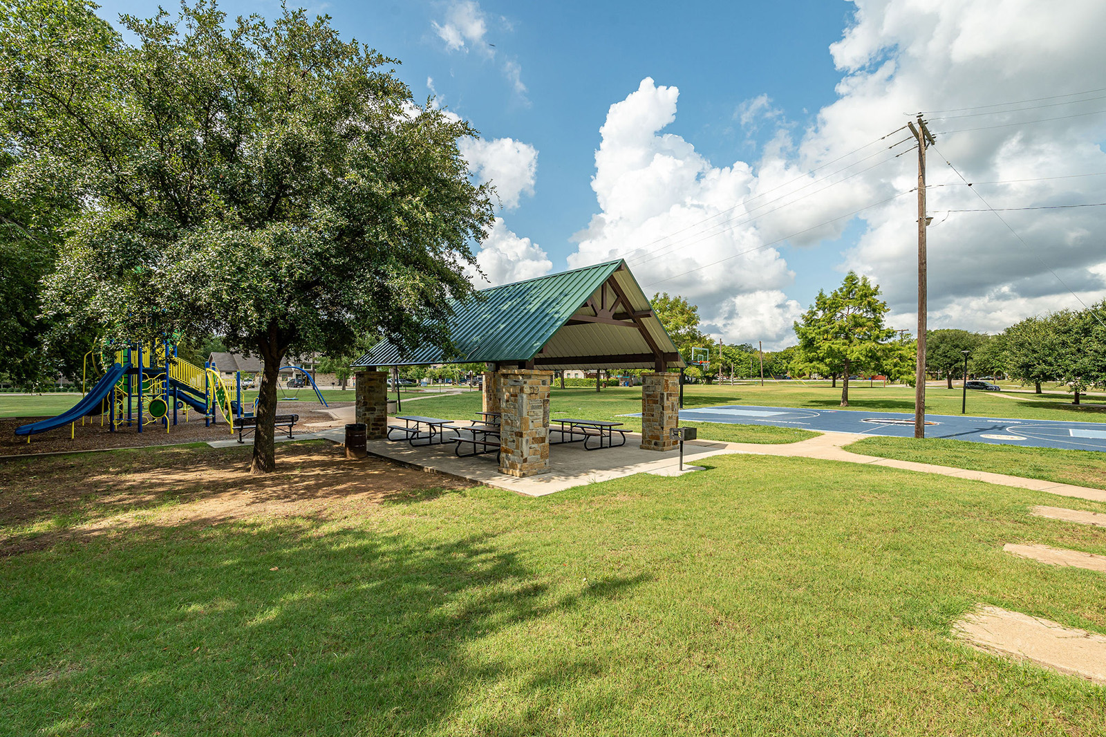 A park with a covered picnic area, playground with slides, basketball court, trees, and grassy open space under a partly cloudy sky.