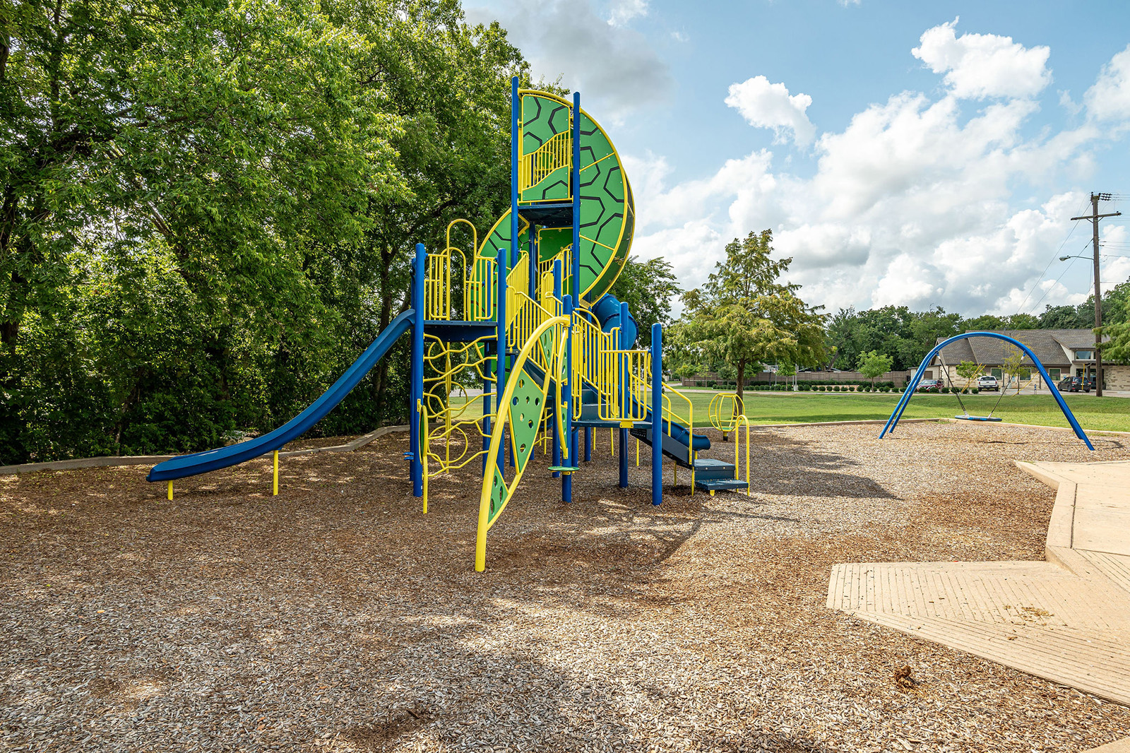 A playground with blue and yellow climbing structures, slides, and monkey bars on a woodchip surface, surrounded by trees and grass under a partly cloudy sky.