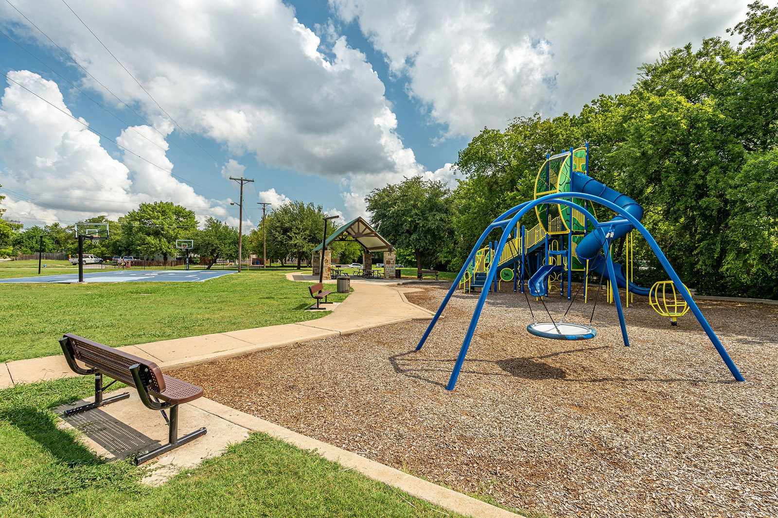 A playground with blue climbing equipment, a swing, a basketball court, benches, and a pavilion set in a grassy park with trees under a partly cloudy sky.