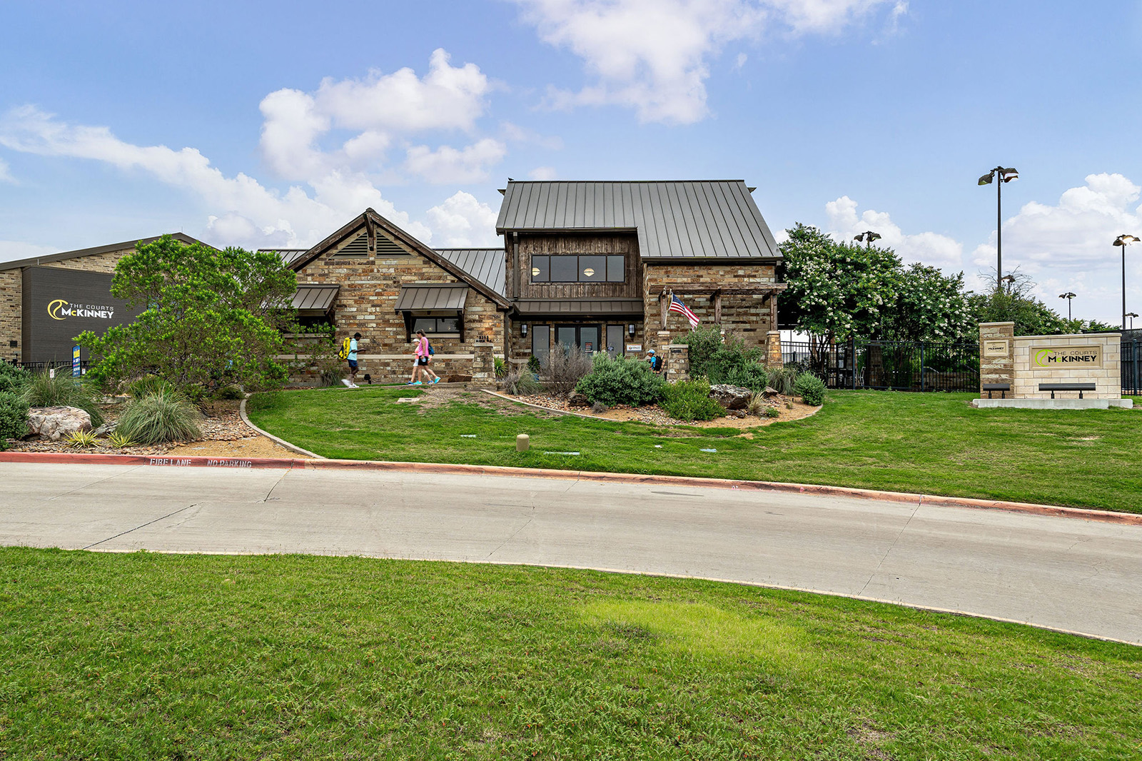 A stone building with a metal roof sits on a well-maintained lawn, with people walking nearby and a sign reading "McKinney" visible on the left and right sides.
