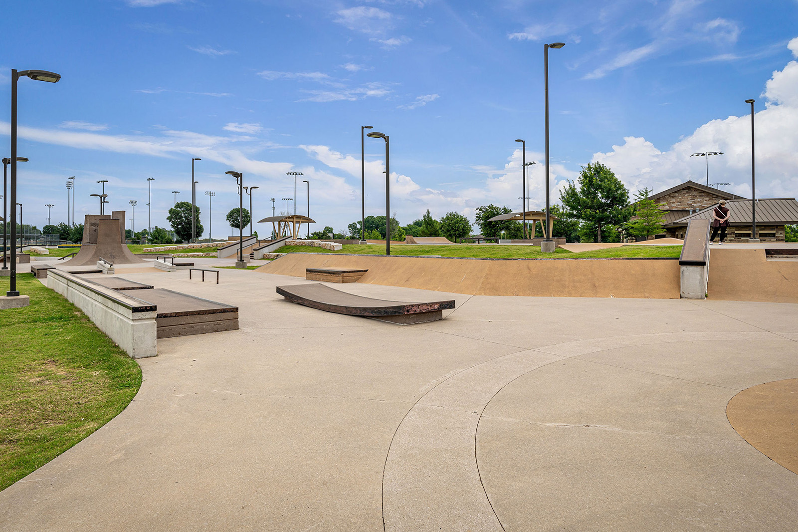 Outdoor skatepark with ramps, rails, and benches under a partly cloudy sky. Surrounding area includes grass, trees, and covered seating structures.