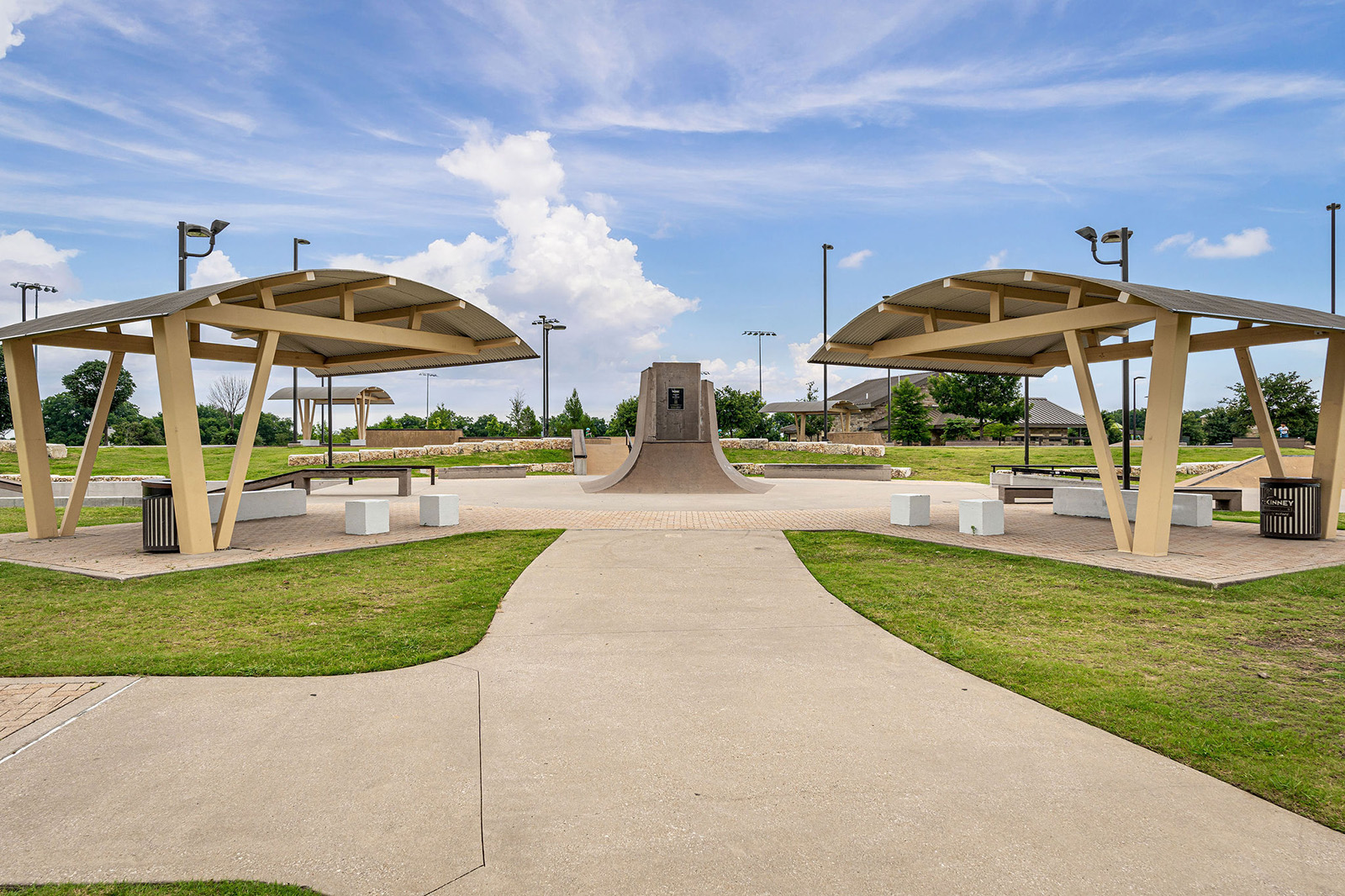 Concrete pathway leading to two covered seating areas with benches and trash cans, and a large skate ramp in the background under a partly cloudy sky.