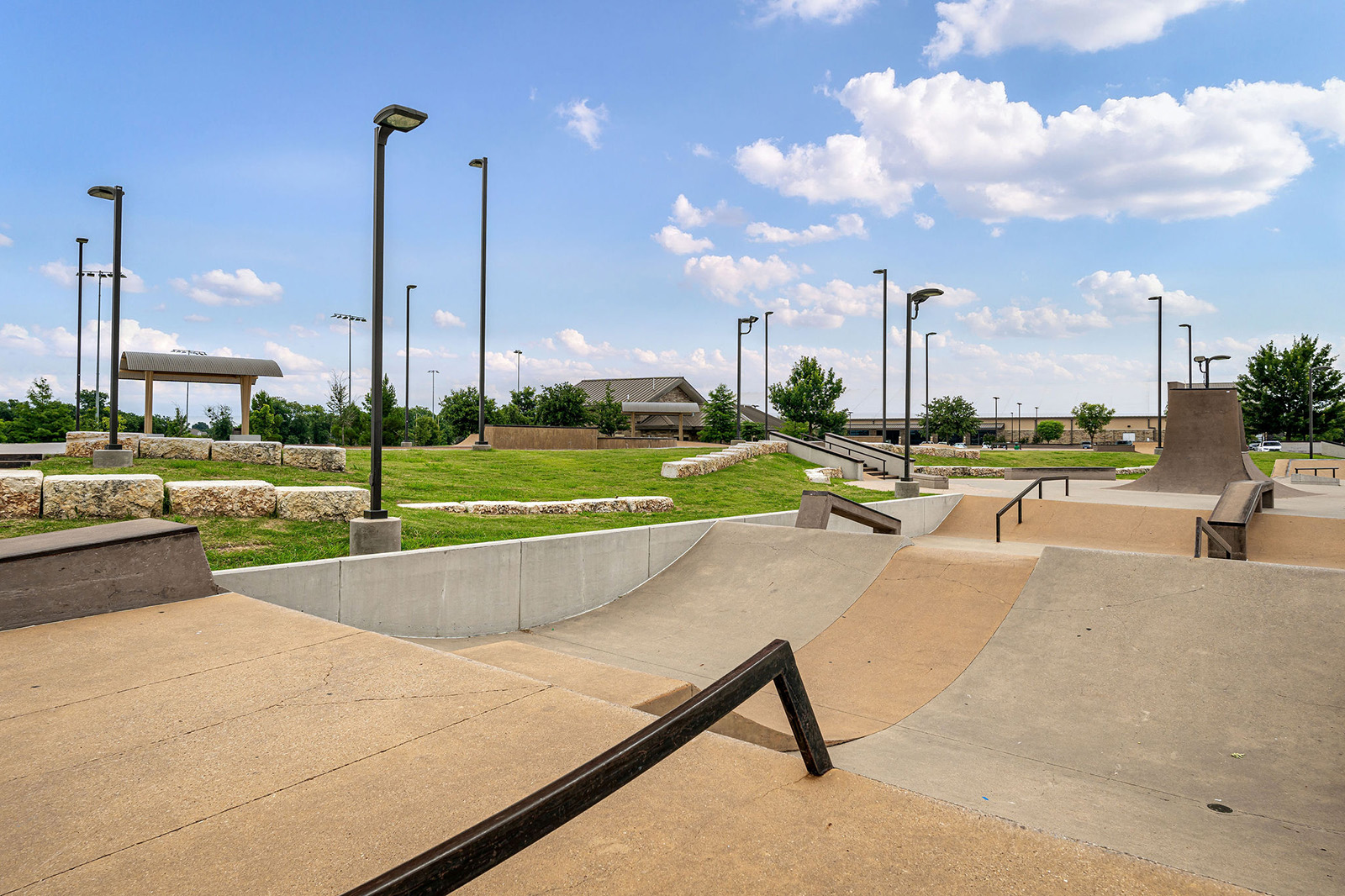 Outdoor skatepark featuring ramps, rails, and ledges, with grassy areas, trees, and light poles visible under a partly cloudy sky.