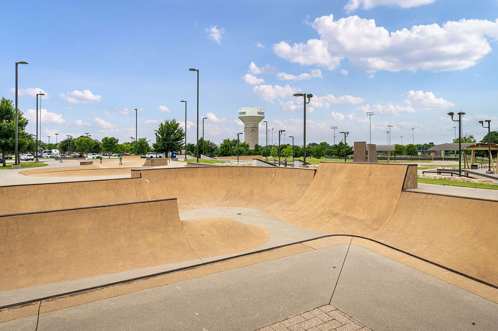 A concrete skatepark with ramps and bowls under a partly cloudy sky, surrounded by trees, light poles, and a distant water tower.