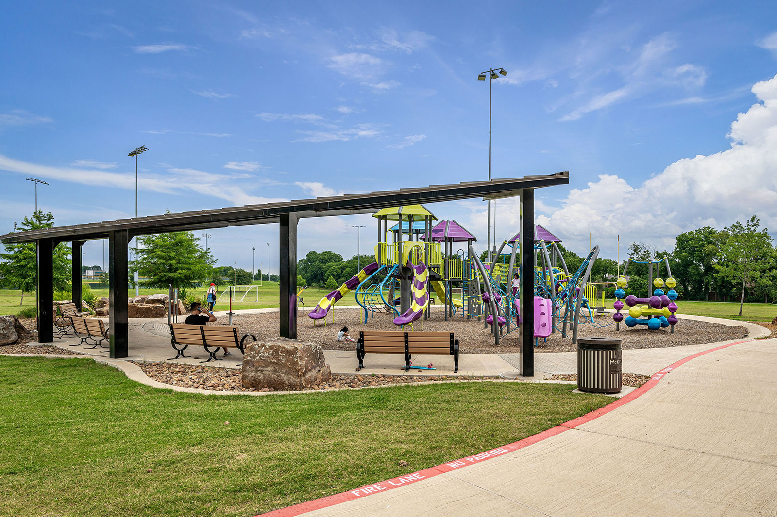 A playground with colorful climbing structures and slides, shaded seating benches, and a trash can on a sunny day in a park.