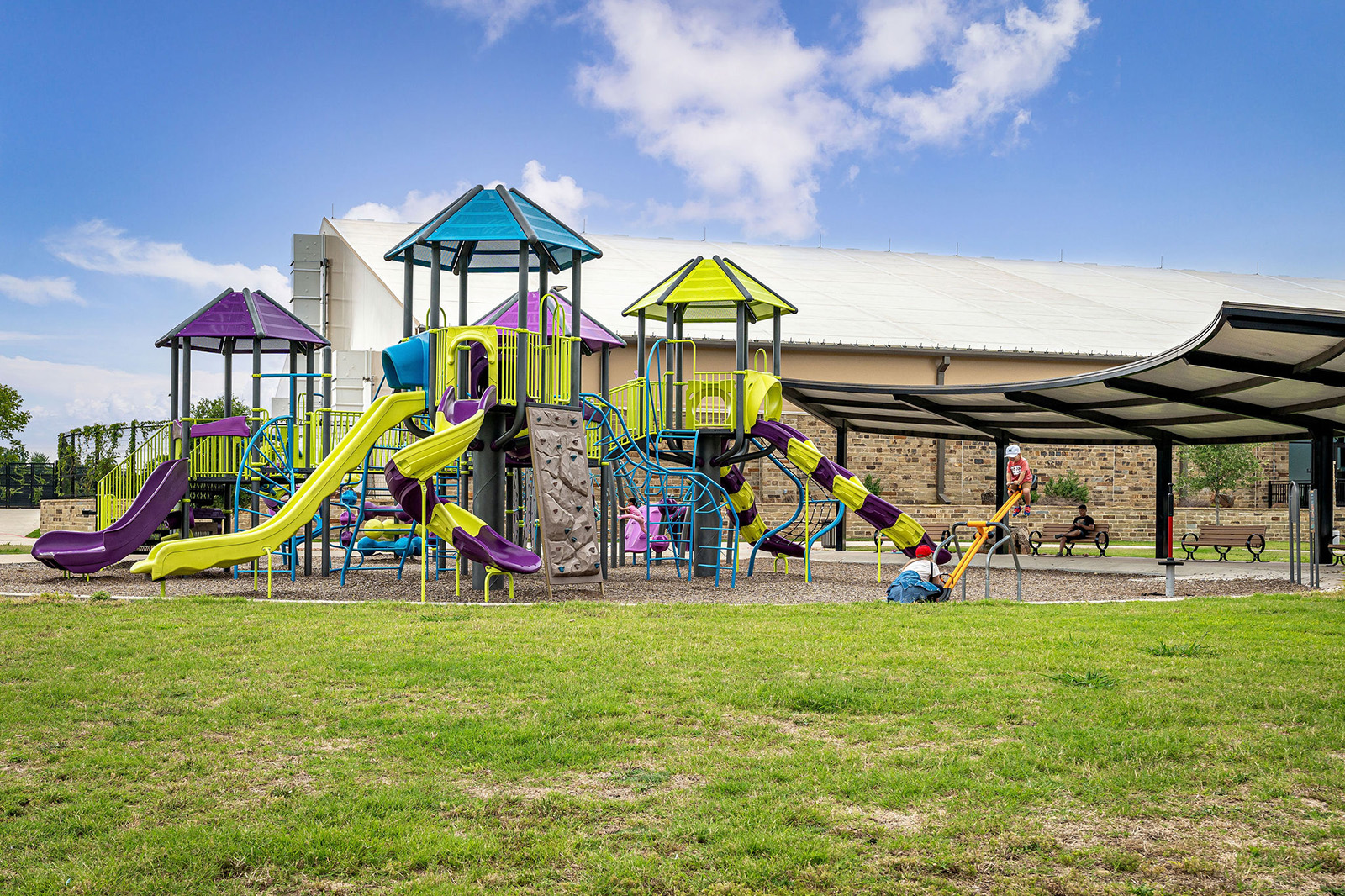 Colorful playground with multiple slides, climbing structures, and children playing. A large building and covered seating area are in the background, with grass in the foreground.
