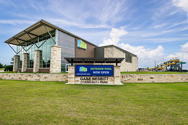 Exterior view of Apex Centre at Gabe Nesbitt Community Park with a sign announcing the outdoor pool is now open; water slides are visible in the background.