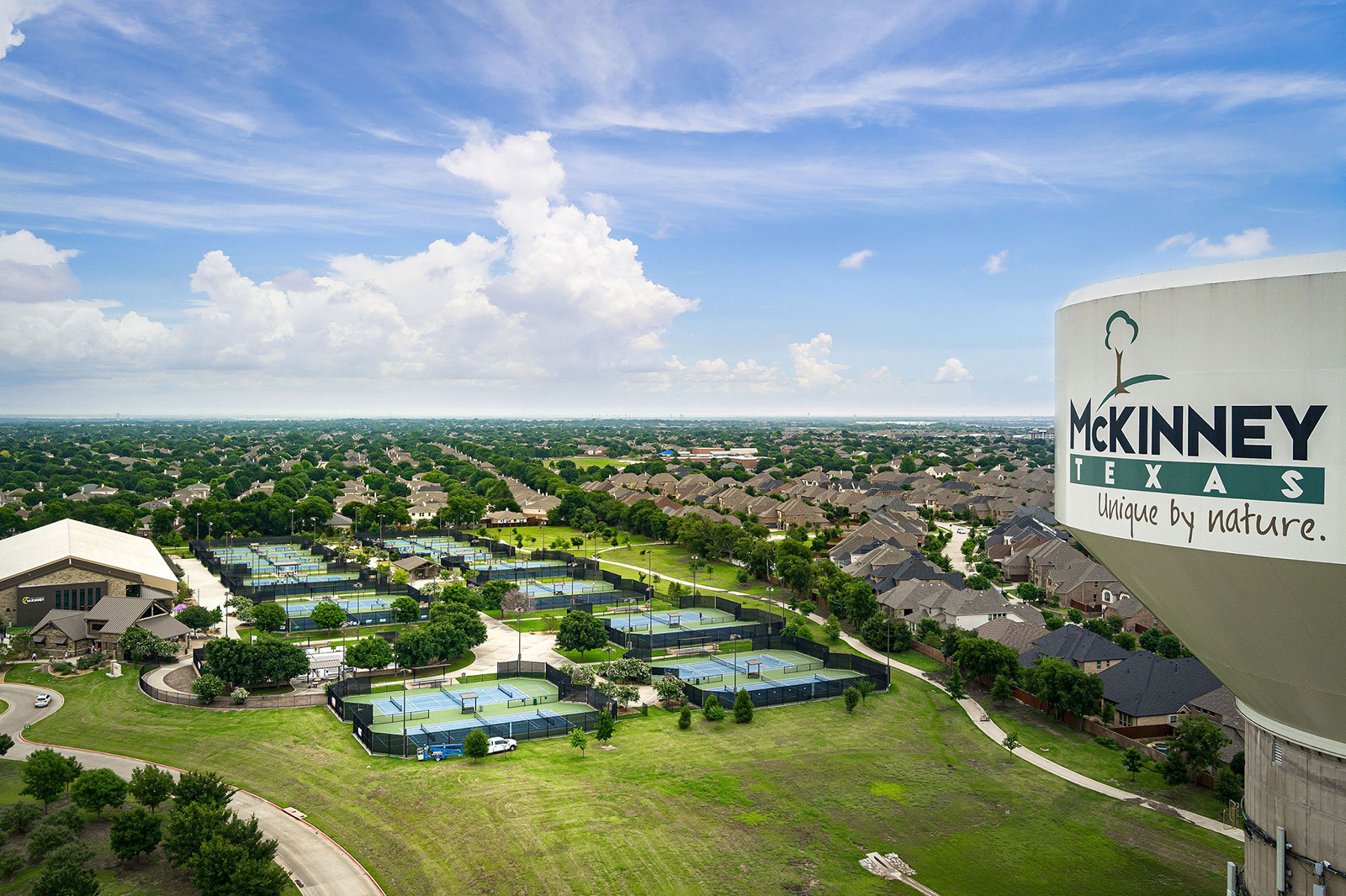 Aerial view of tennis courts, residential houses, and a large water tower labeled "McKinney Texas" in a suburban area under a partly cloudy sky.