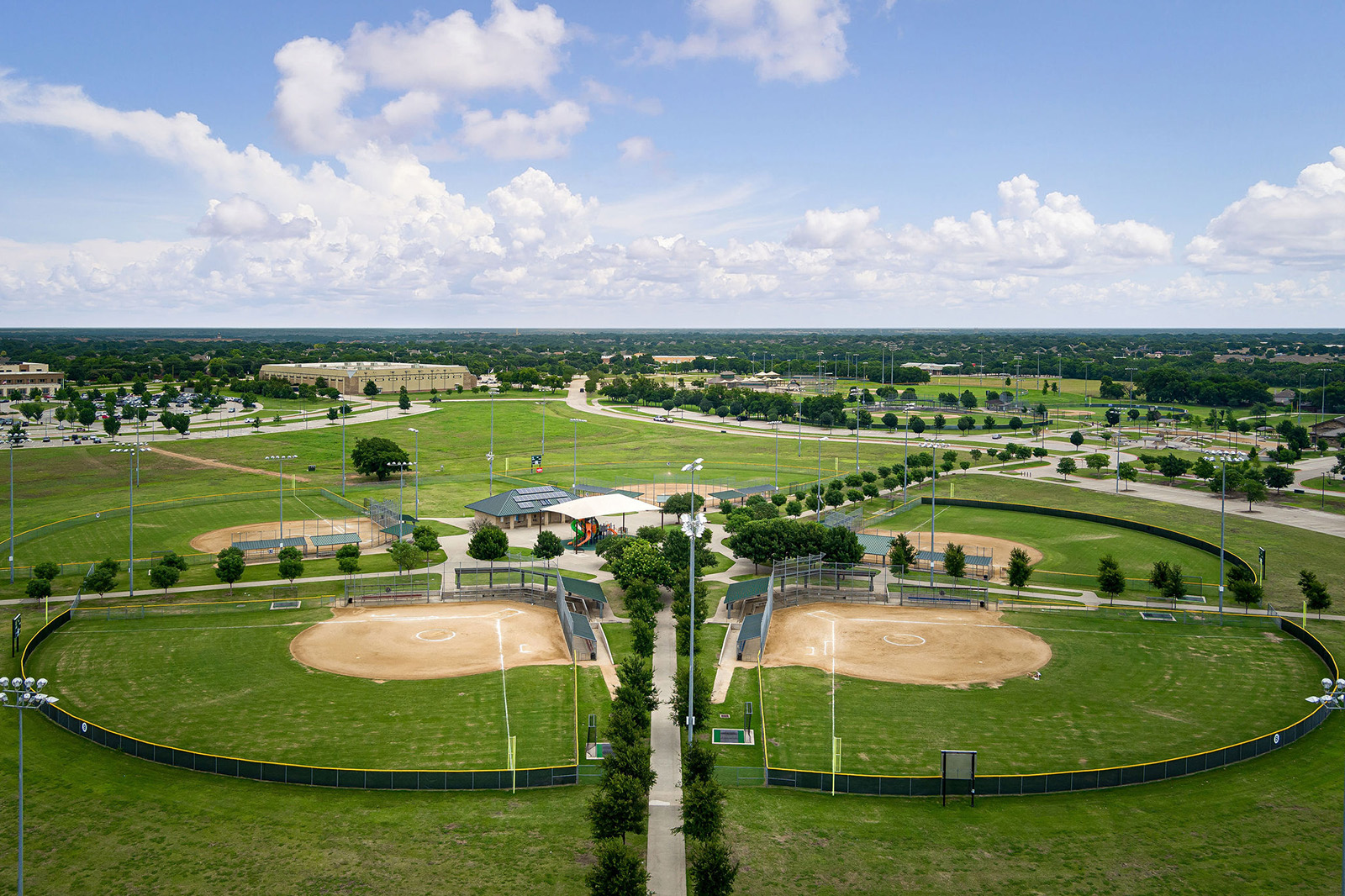 Aerial view of a sports complex featuring two adjacent baseball or softball fields with green grass, dirt infields, and surrounding pathways under a partly cloudy sky.