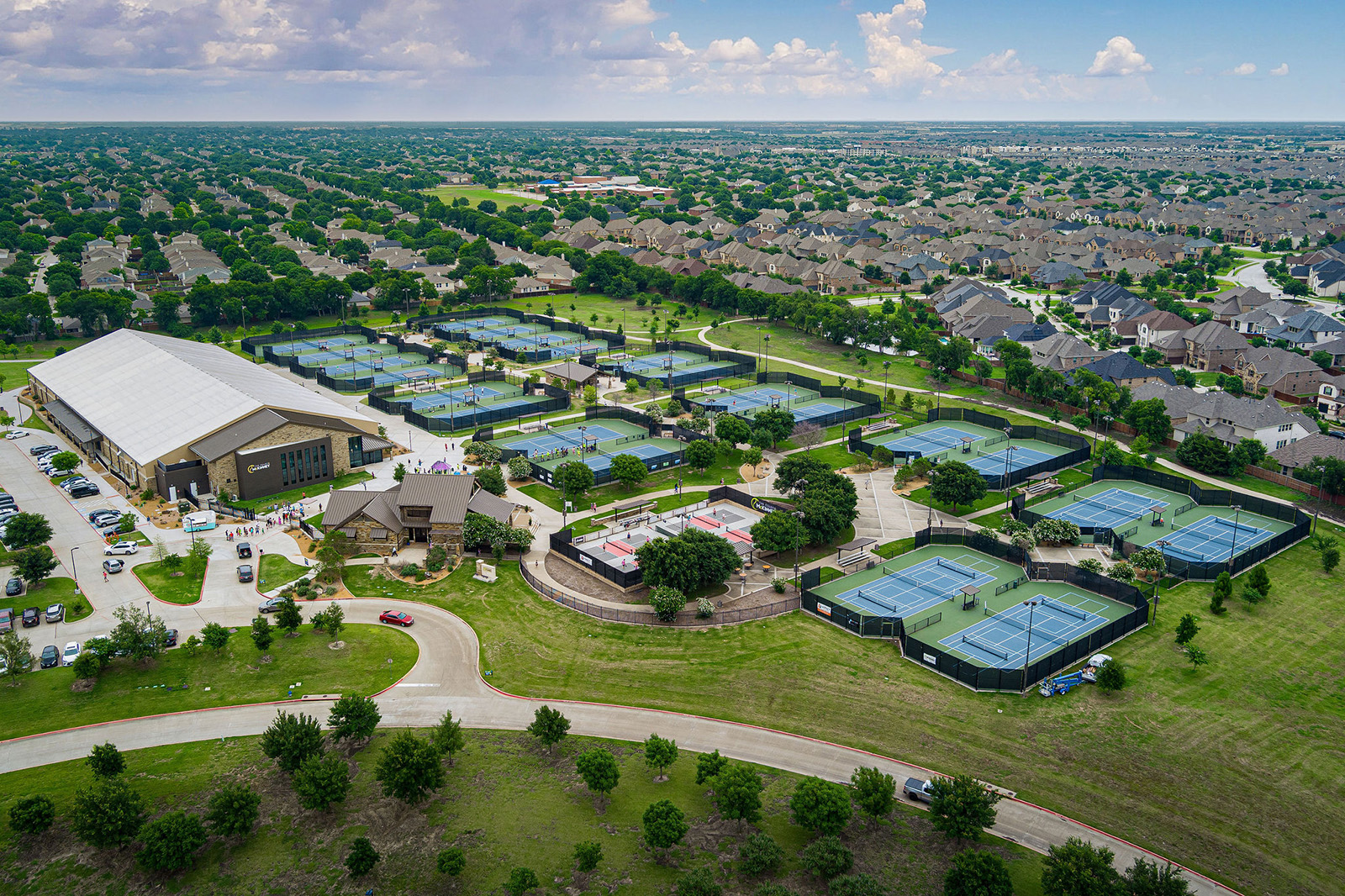 Aerial view of a large tennis complex with multiple outdoor courts, an indoor facility, parking lots, and surrounding residential neighborhoods.