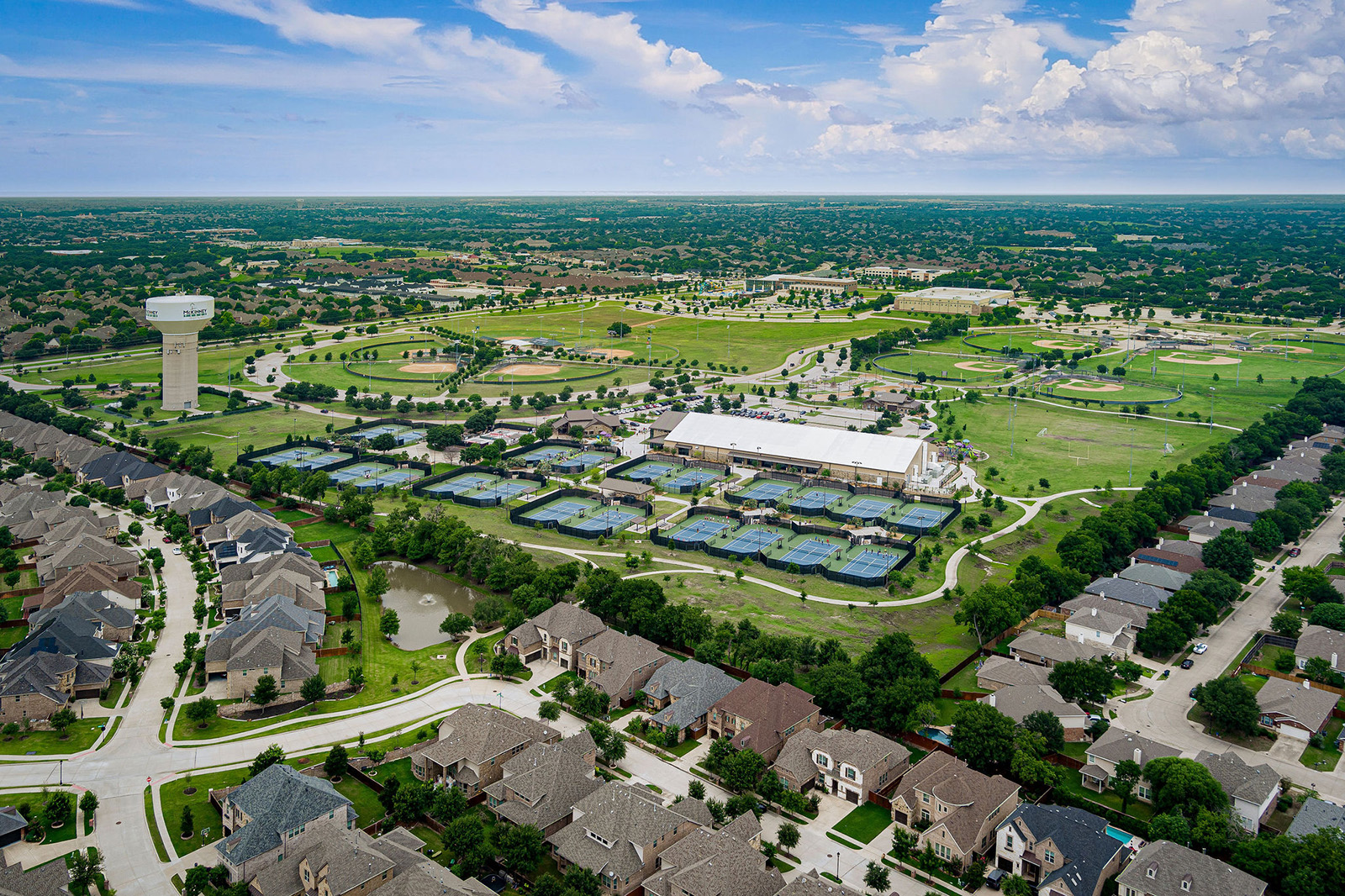 Aerial view of a suburban neighborhood with rows of houses, a large water tower, and multiple tennis courts near a recreational facility surrounded by green fields.
