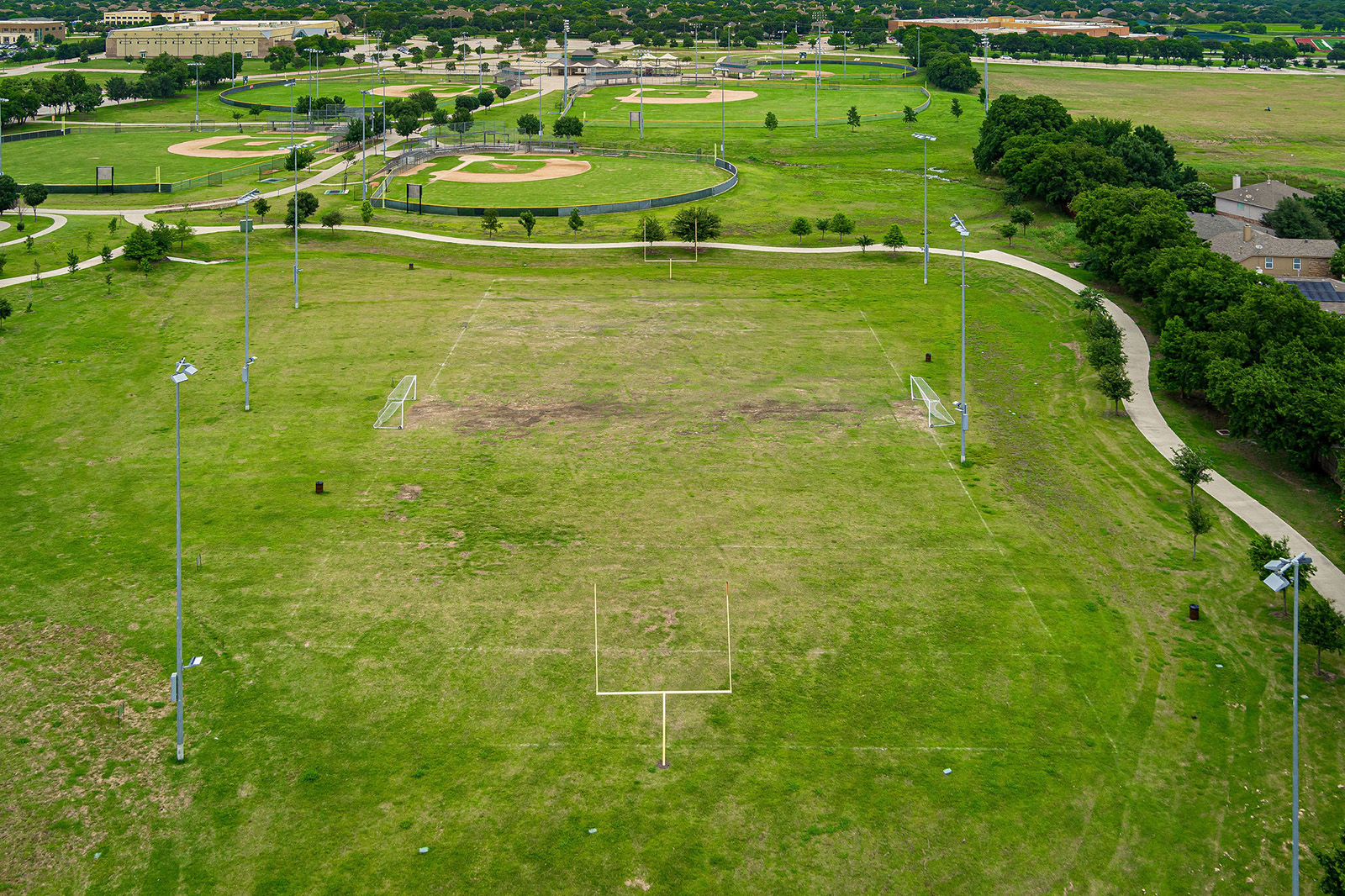 Aerial view of an empty grass sports field with goalposts, surrounded by light poles, pathways, and baseball fields in the background.
