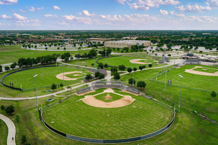 Aerial view of a sports complex featuring multiple baseball fields surrounded by green grass, trees, and parking lots under a partly cloudy sky.