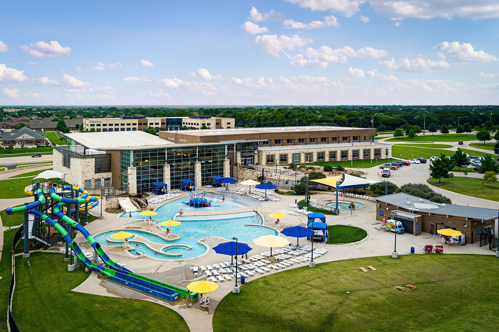 Aerial view of an outdoor water park with slides, pools, lounge chairs, yellow umbrellas, and a large modern building in the background on a sunny day.