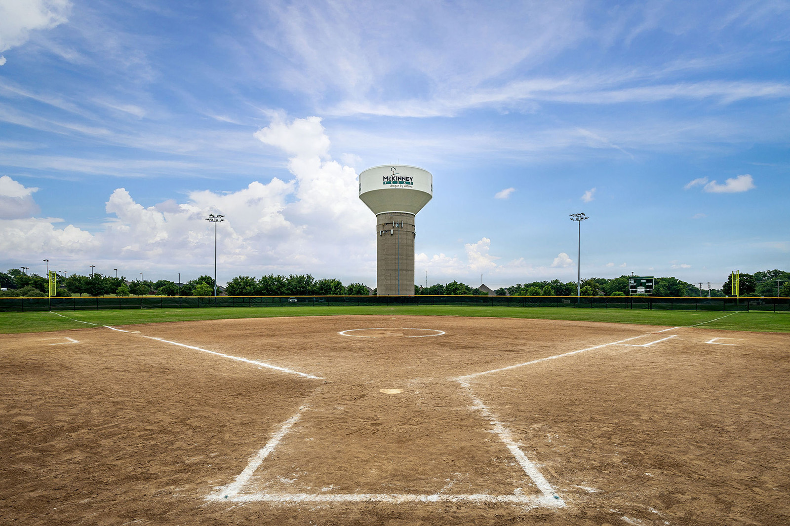 Empty baseball field with a dirt infield and white baselines under a blue sky, with a large water tower in the background marked "The Colony.