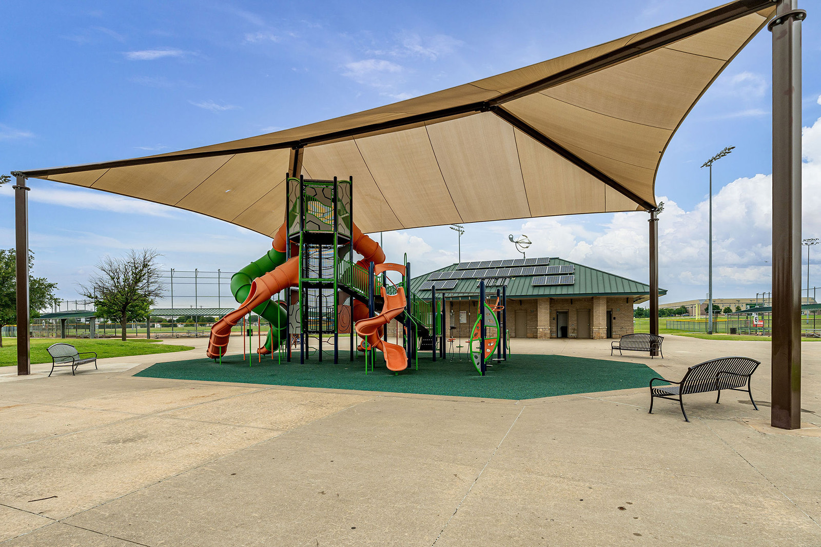 A modern playground with orange and green slides sits under a large shade canopy next to a building and benches on a concrete surface.