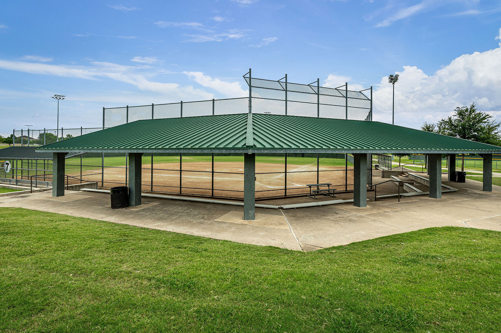 A covered outdoor baseball field with a green metal roof, concrete walkway, benches, and protective fencing under a partly cloudy sky.