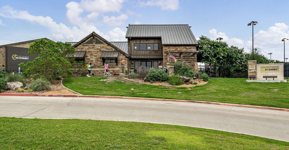A stone and wood building with a metal roof sits behind a landscaped lawn. Several people walk nearby, and a sign reads "McKinney" by the entrance.
