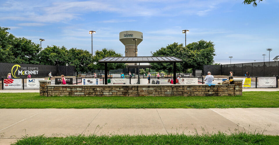 A covered seating area overlooks multiple tennis courts at The Courts of McKinney, with people watching and playing under a partly cloudy sky.
