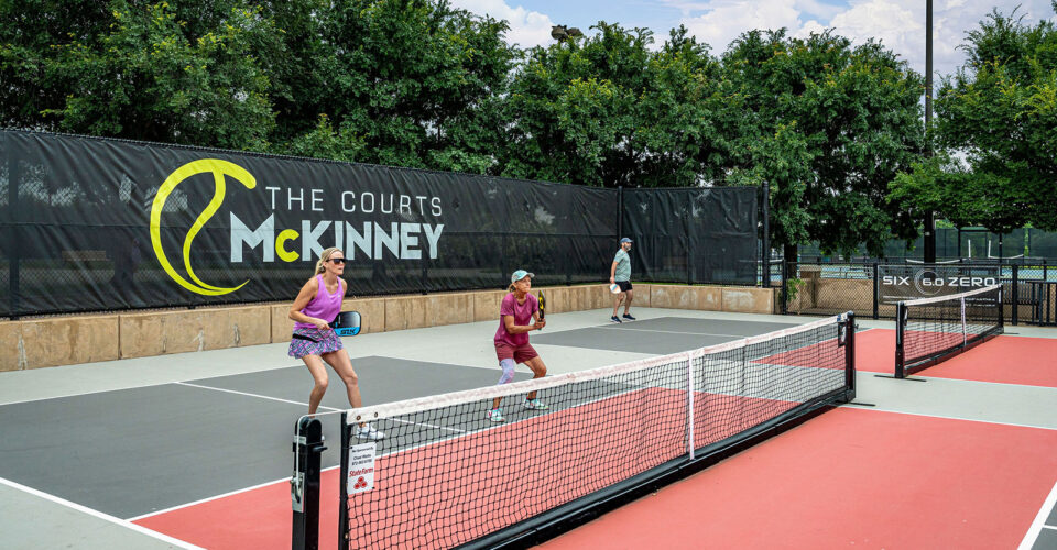 Two pairs of people play pickleball on outdoor courts at The Courts McKinney, with trees and a black fence in the background.