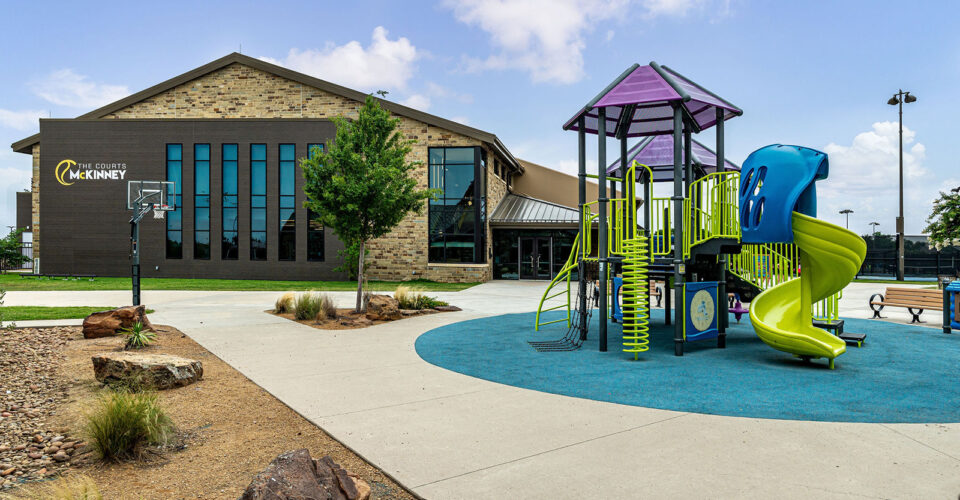 A modern playground with a slide and climbing structure sits in front of a stone building labeled “City of McKinney” under a partly cloudy sky.