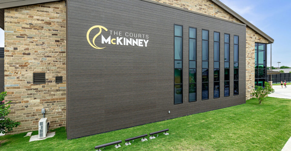 Exterior view of The Courts McKinney building with large windows, stone and dark siding, and grass lawn under a clear sky.